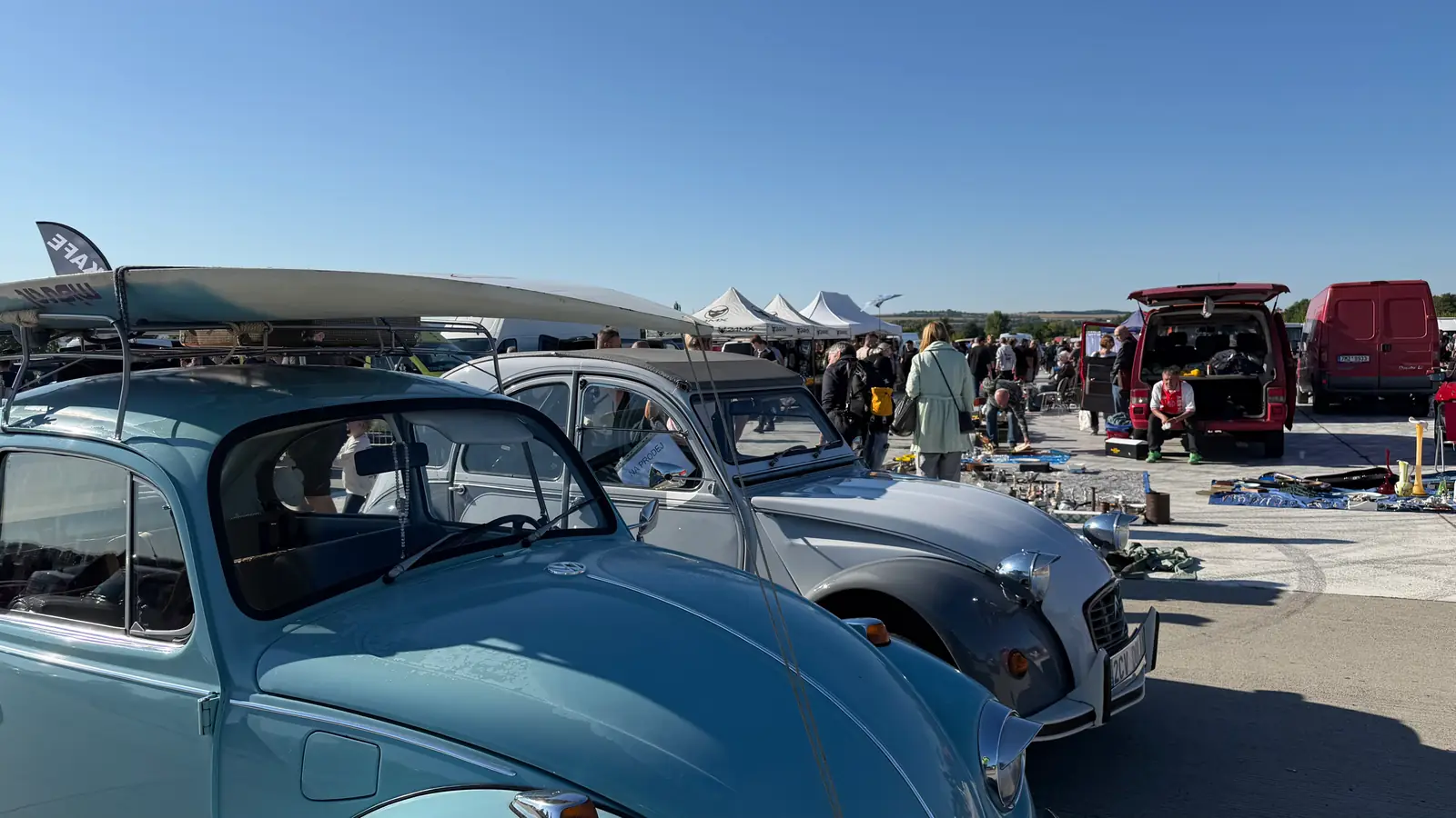 Světle modrý VW Brouk a Citroën 2CV stojí v řadě, za nimi stany prodejců